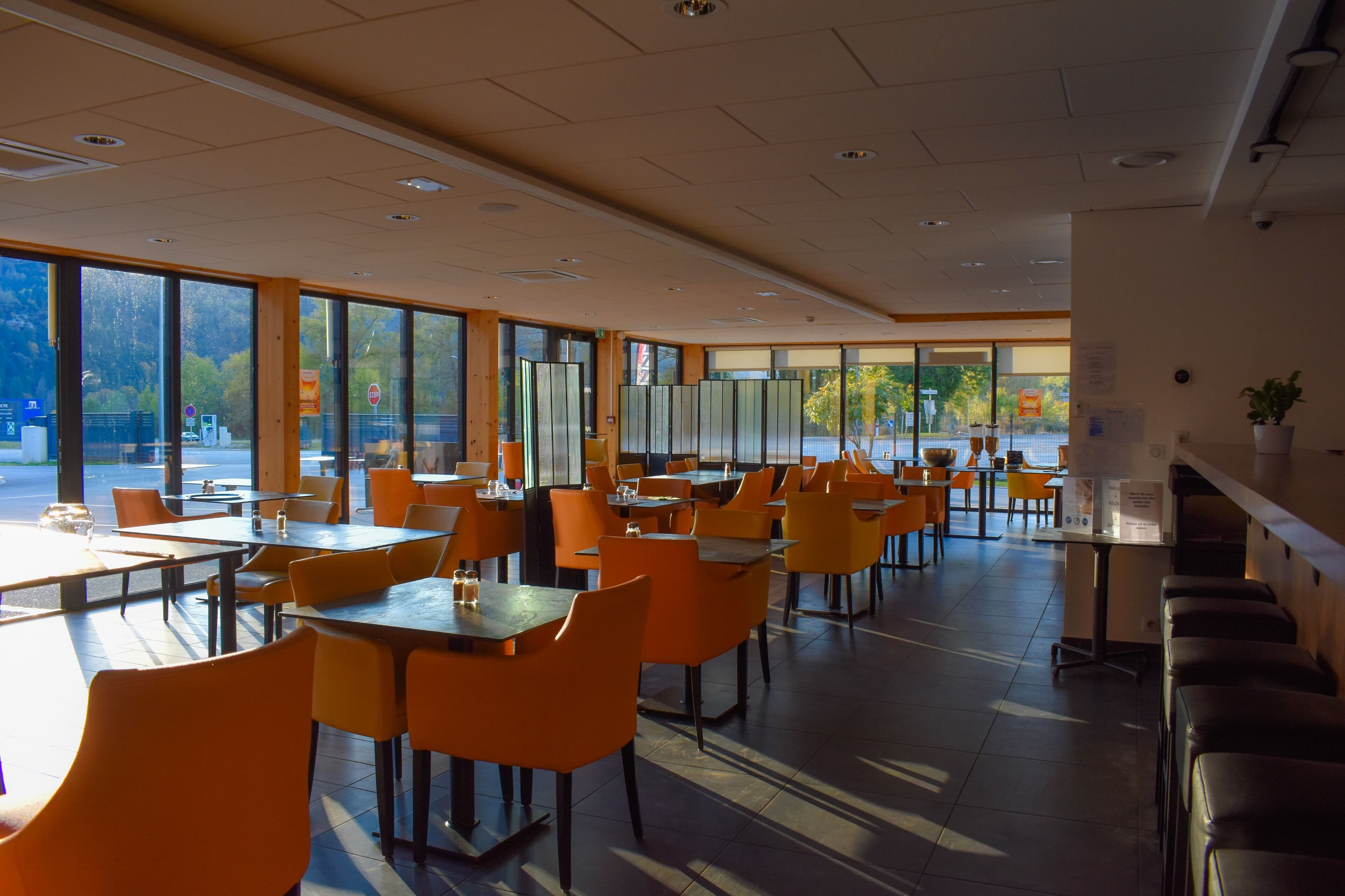 A spacious restaurant with high tables and orange chairs, large windows offering views of the outdoors, and a clean, modern interior.