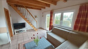 A living room with a wooden staircase, a television on a white stand, a beige sofa, and a large window with curtains.