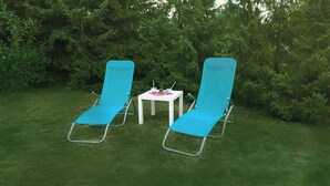 Two blue lawn chairs with metal frames and a small white table with a pink placemat, surrounded by green grass and dense foliage.