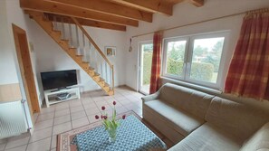 A living room with a wooden staircase, a television on a white stand, a beige sofa, and a large window with curtains. - Vacation home "Jessica" in country house style near national park beautiful bike paths and beaches  (Rechlin)