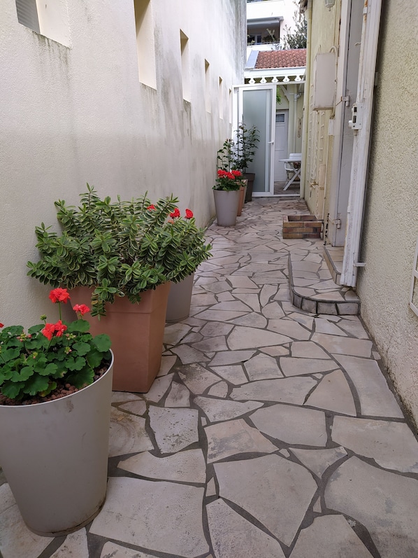 Um corredor estreito com piso de pedra, vasos de plantas e uma porta que dá para uma área coberta.