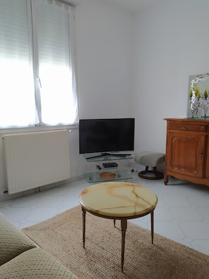 Sala de estar com televisão de tela plana em suporte de vidro, um armário de madeira, uma mesa redonda e um radiador.