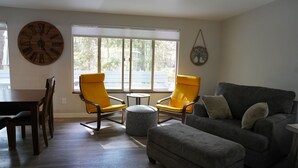 A living room with a wooden floor, two yellow armchairs, a grey sofa, a round ottoman, and a large wall clock. - Updated 2bd/2ba. Walk to Village, dog friendly, SHARC passes. (Sunriver)