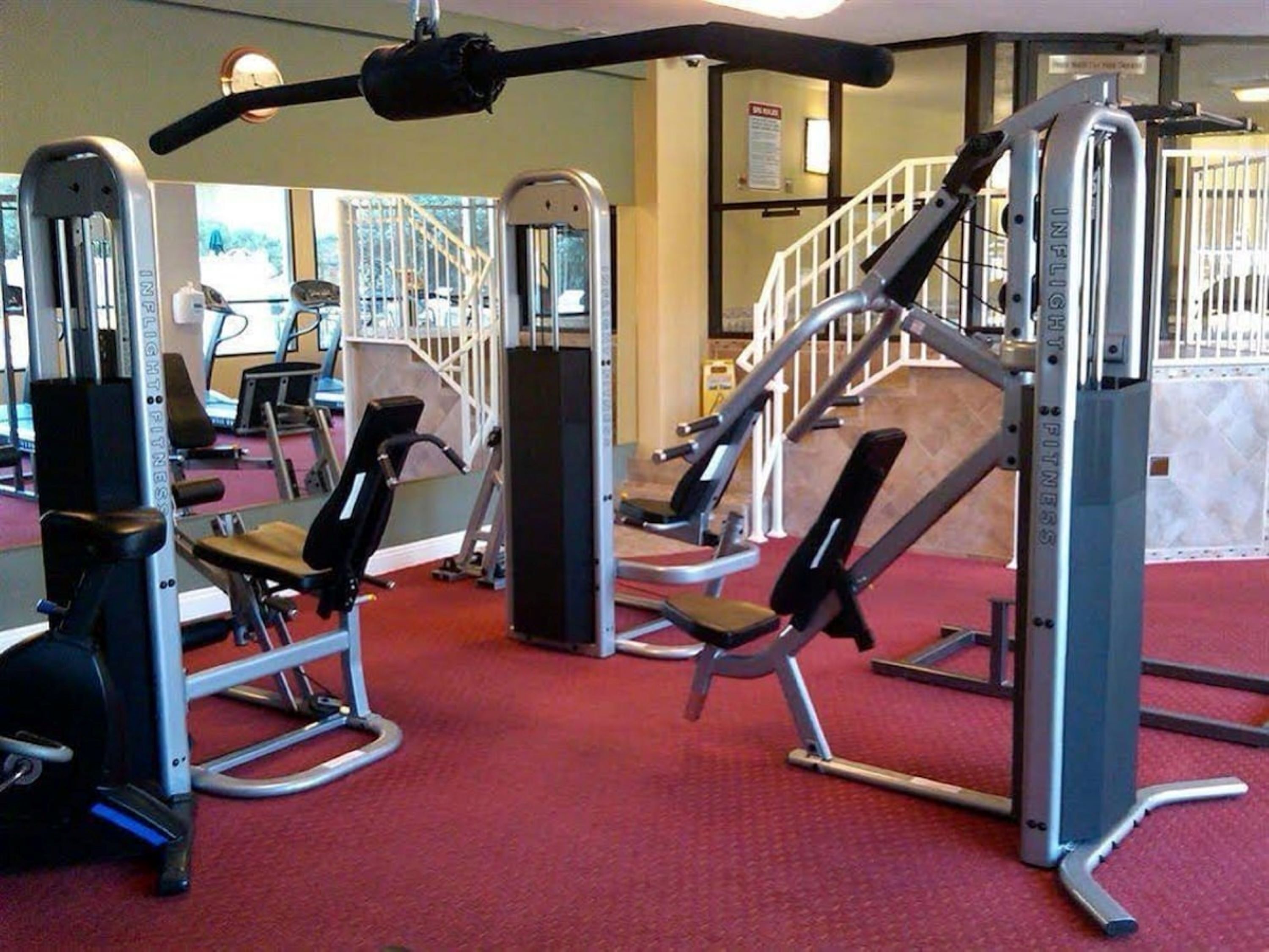 Un gimnasio con diversas máquinas de ejercicio, piso de moqueta roja y una escalera al fondo.