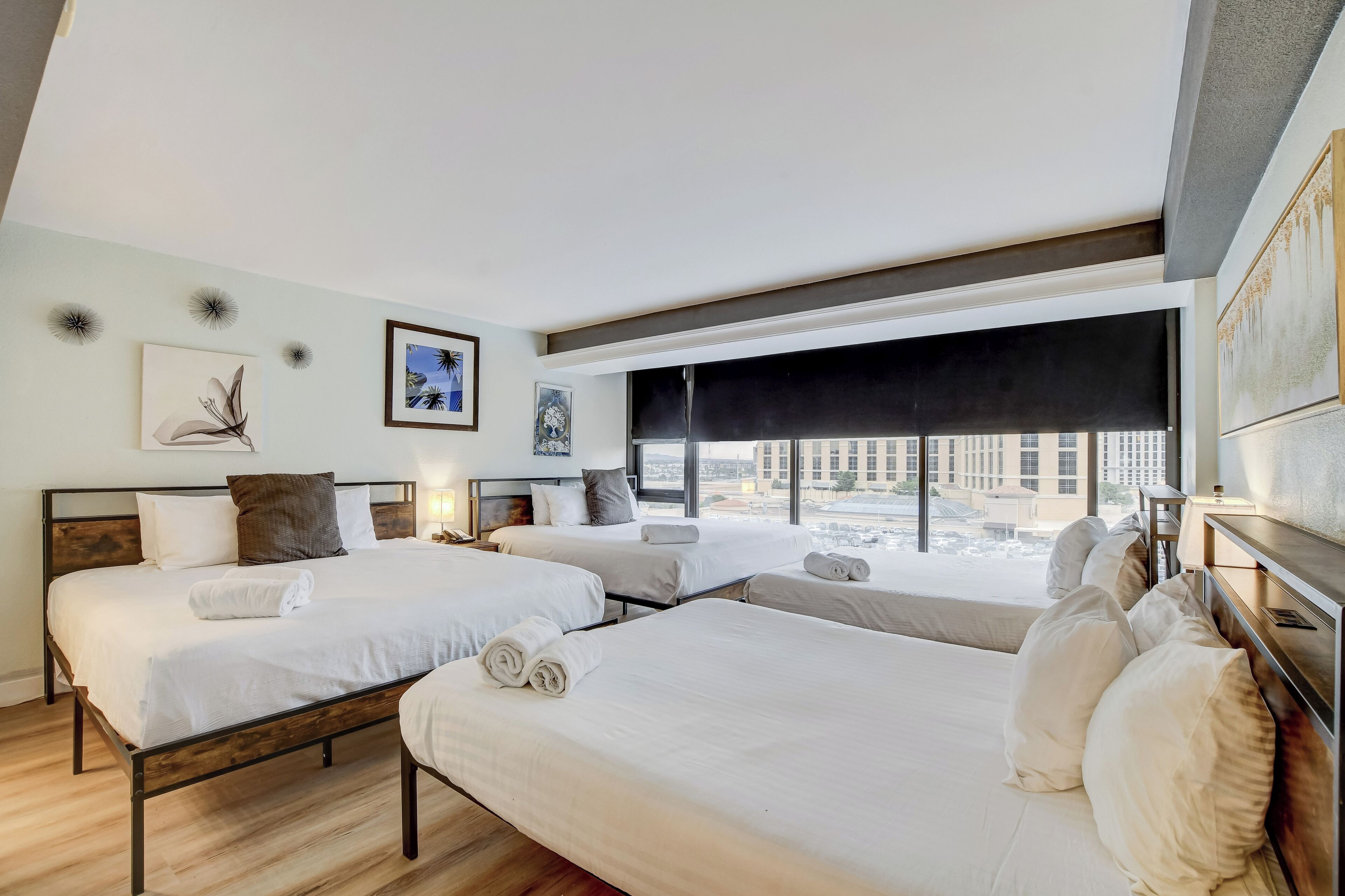 Quarto de hotel com três camas, janelas amplas e obras de arte nas paredes.