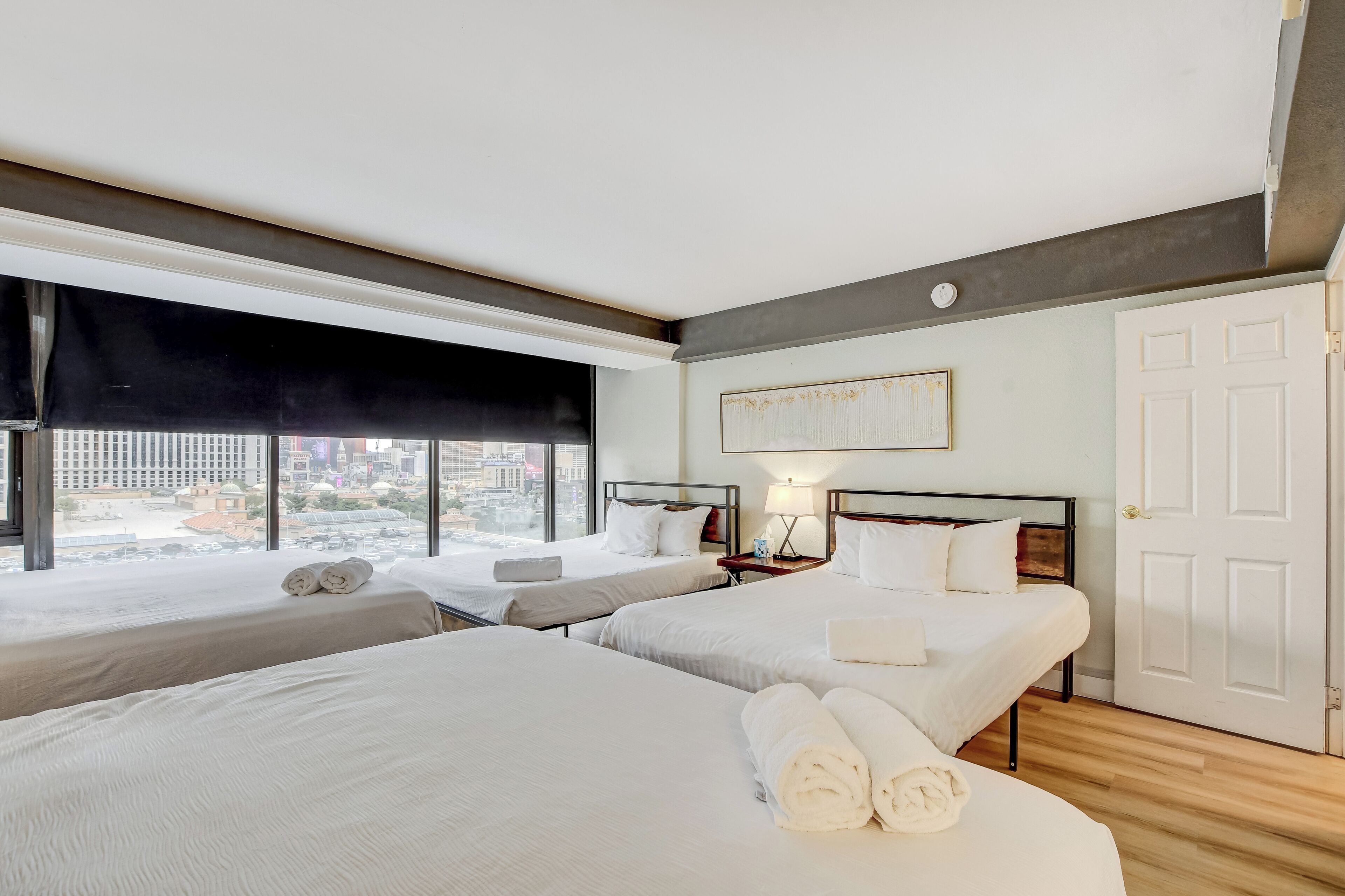 A hotel room with two beds, a large window with a black blind, a bedside table with a lamp, and a door.