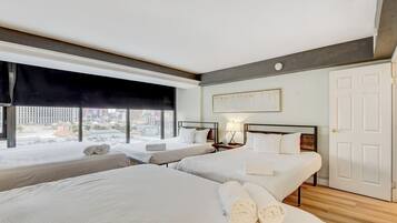 A hotel room with two beds, a large window with a black blind, a bedside table with a lamp, and a door.