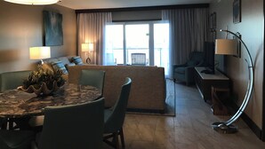A modern hotel room with a dining area, a sofa, a TV, and a large window with curtains.