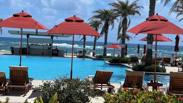 A poolside area with red umbrellas, lounge chairs, and a bar with a view of the ocean.