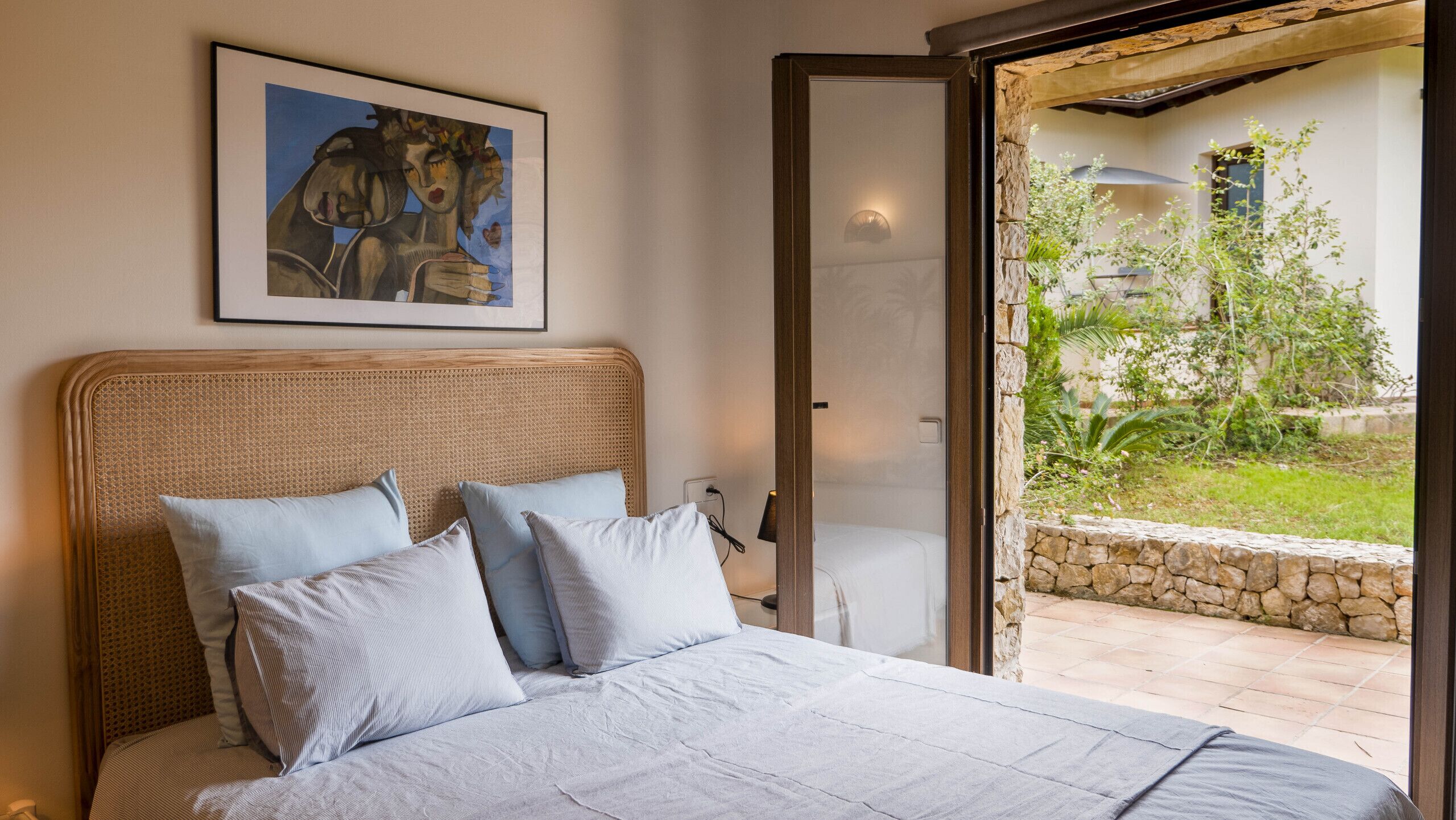 A bedroom with a large bed, a rattan headboard, and a view of a garden through an open door.