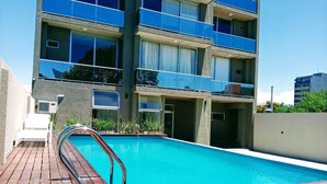 A modern building with a swimming pool, wooden deck, and a white chair. - DFC COLIVING (Salta)