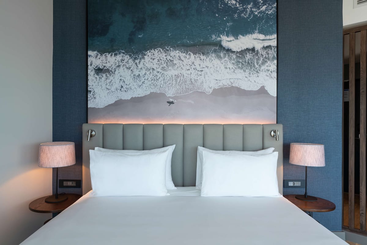 A hotel room with a large bed, two bedside tables with lamps, a grey upholstered headboard, and a large wall mural of ocean waves.