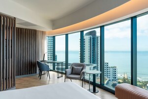 A modern hotel room with a large window, a bed, a small table, two chairs, and a view of the ocean and buildings.