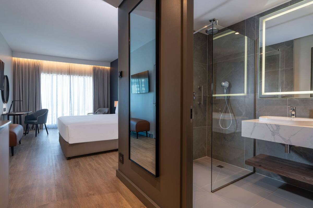 A modern hotel room with a large bed, a desk, a chair, a TV, a shower area, and a bathroom with a mirror and sink.