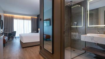 Una habitación de hotel moderna con una cama grande, un escritorio, una silla, un televisor, zona de ducha y un baño con espejo y lavamanos.