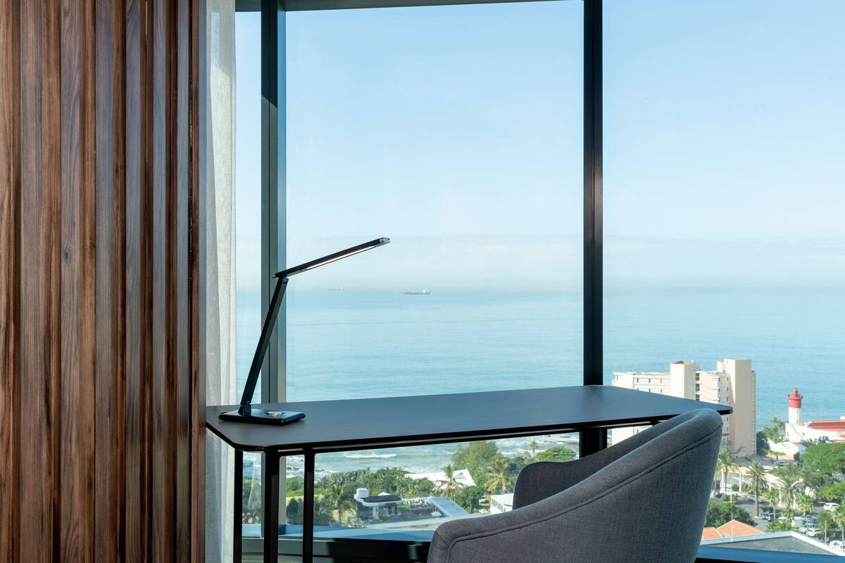 A modern room with a large window overlooking the sea, a desk with a lamp, and a comfortable chair.