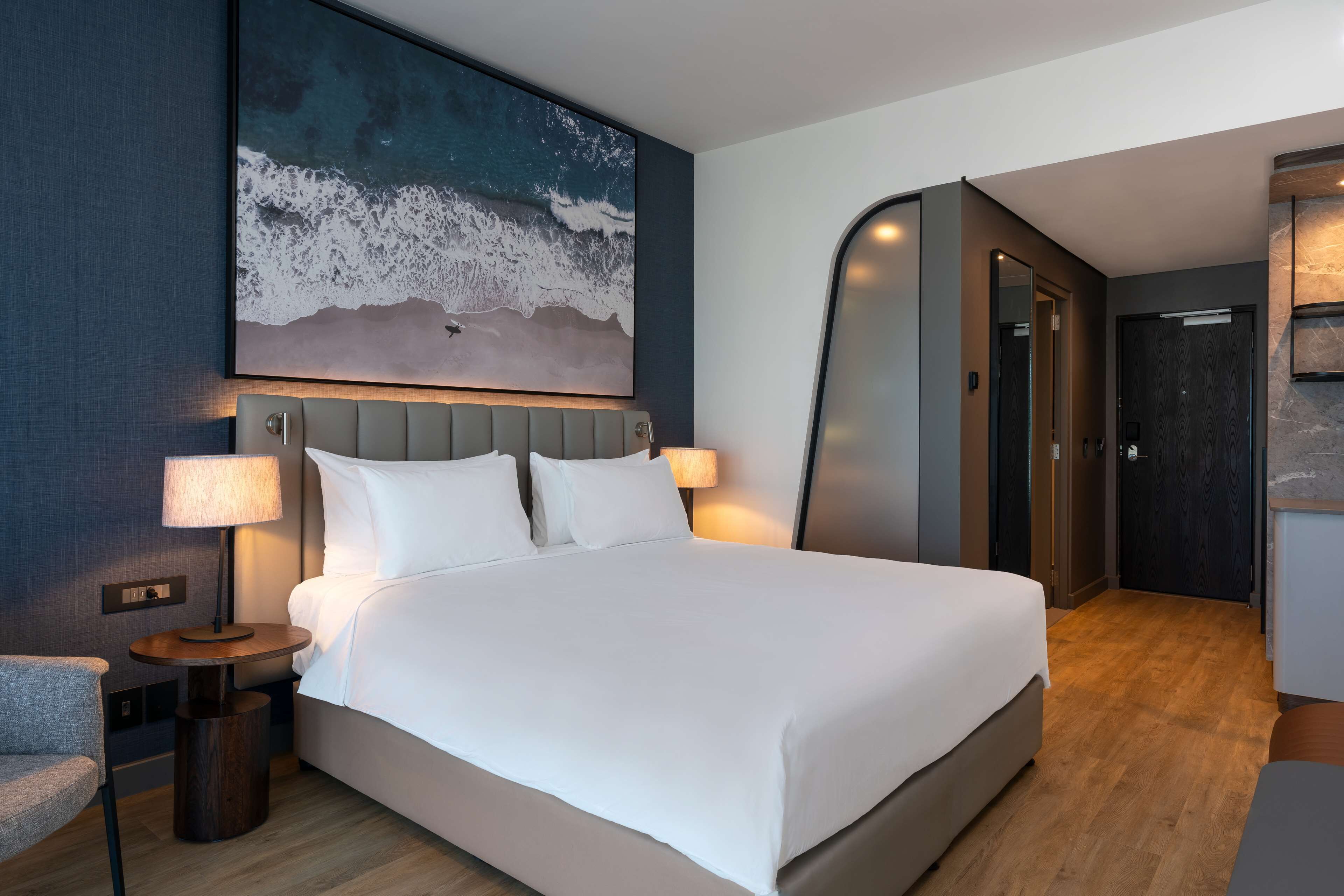 A modern hotel room with a large bed, two bedside lamps, a headboard, and a wall-mounted artwork of ocean waves.