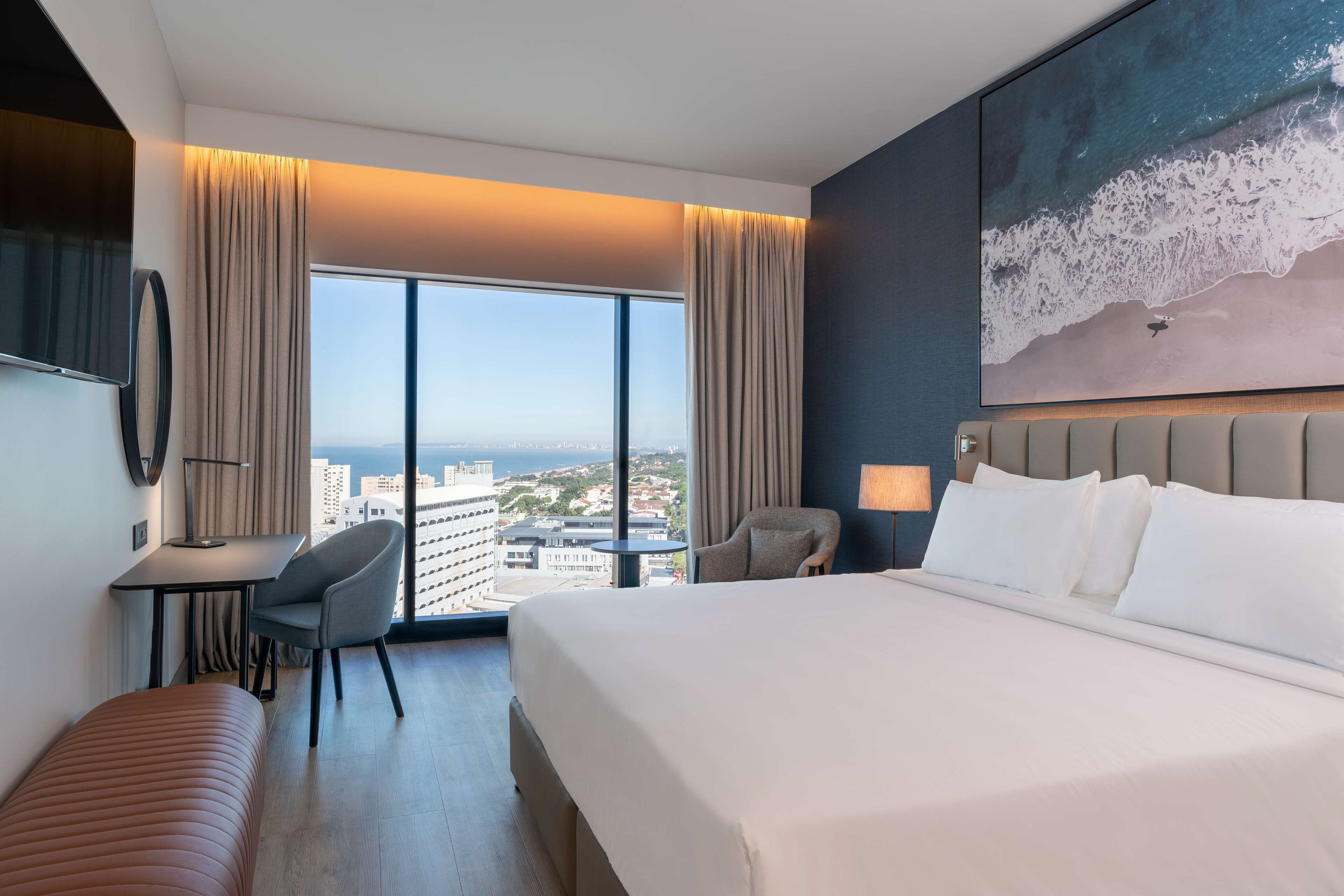 A hotel room with a large bed, a desk, a chair, and a view of the cityscape.