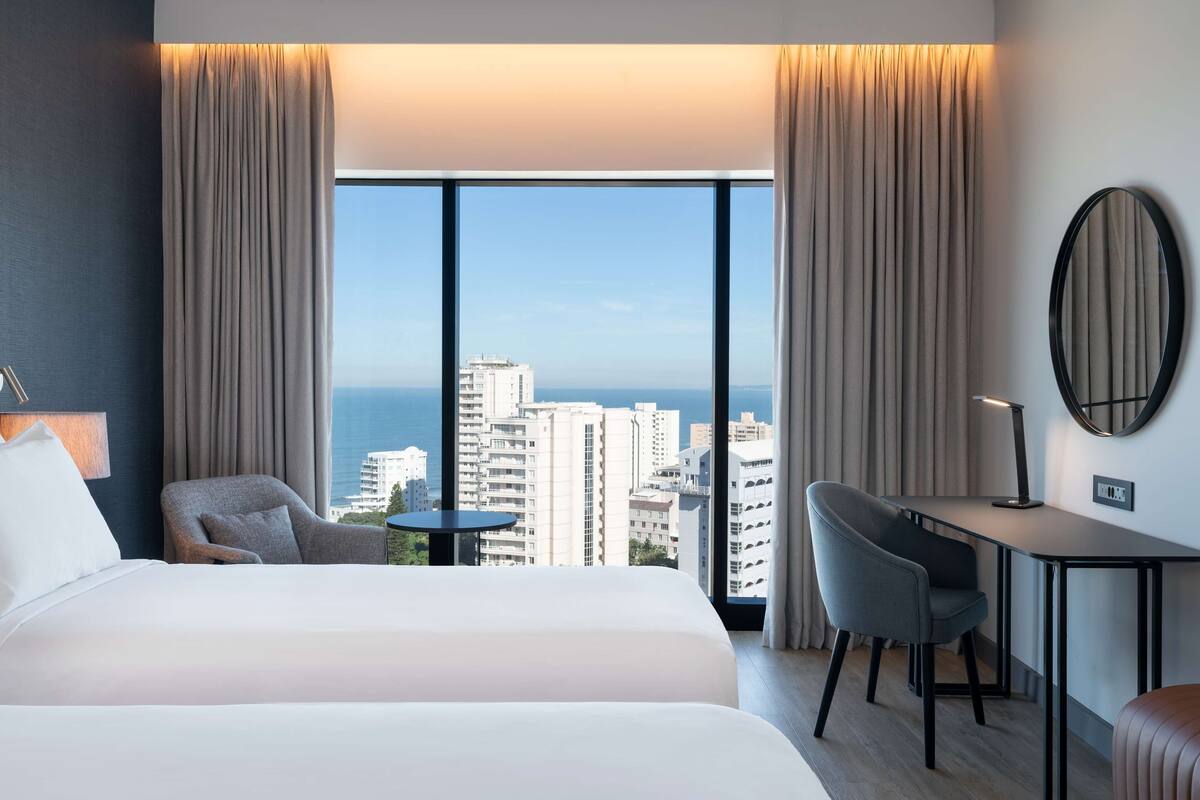 A hotel room with a large bed, a desk, a chair, and a view of the cityscape through large windows.