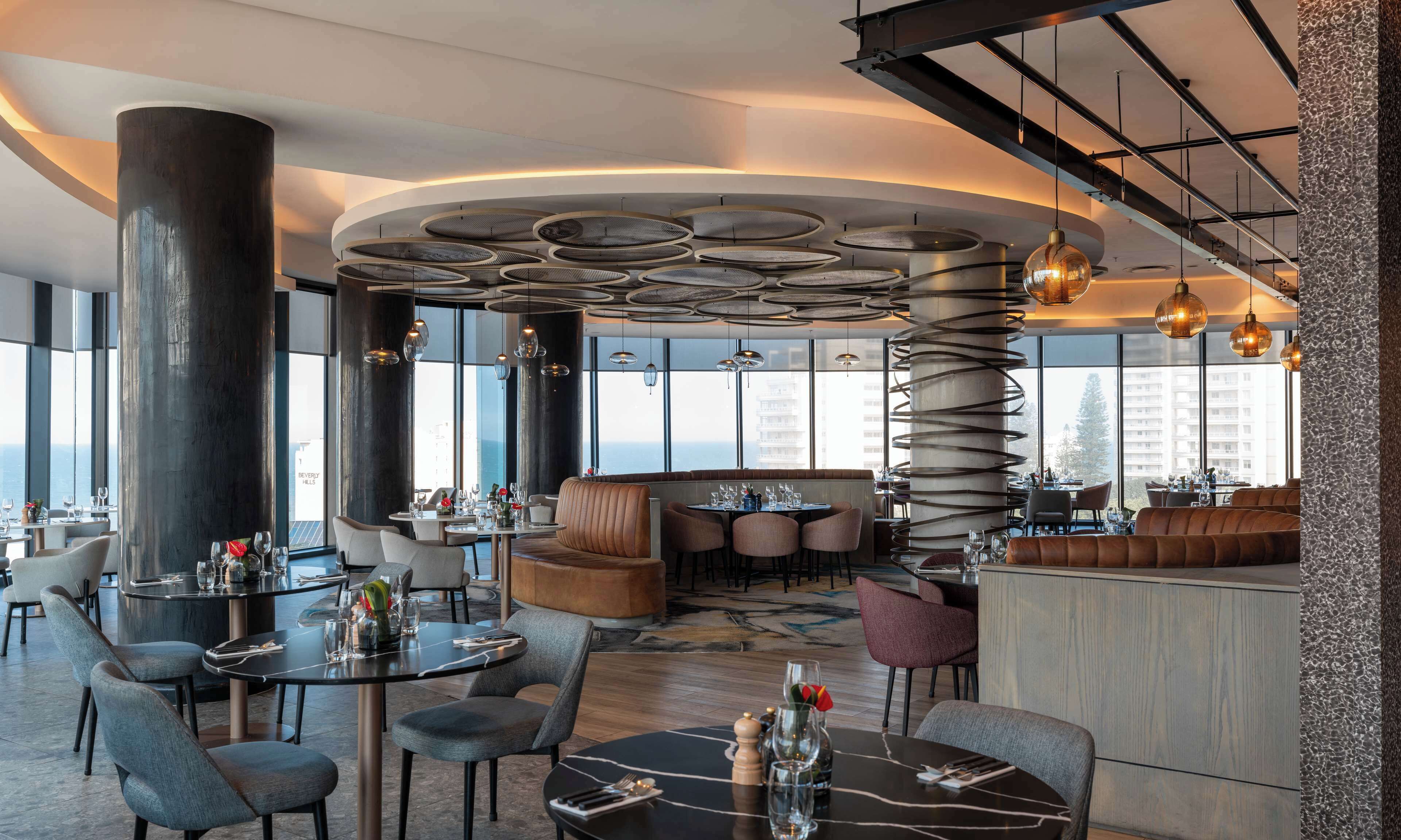 A modern restaurant with round tables, high ceilings, and large windows offering views of the city.