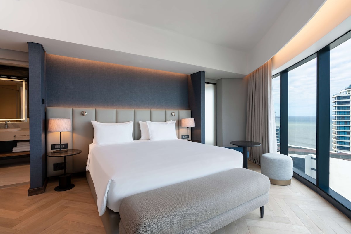 A modern hotel room with a large bed, bedside lamps, a round table, and a view of the sea.
