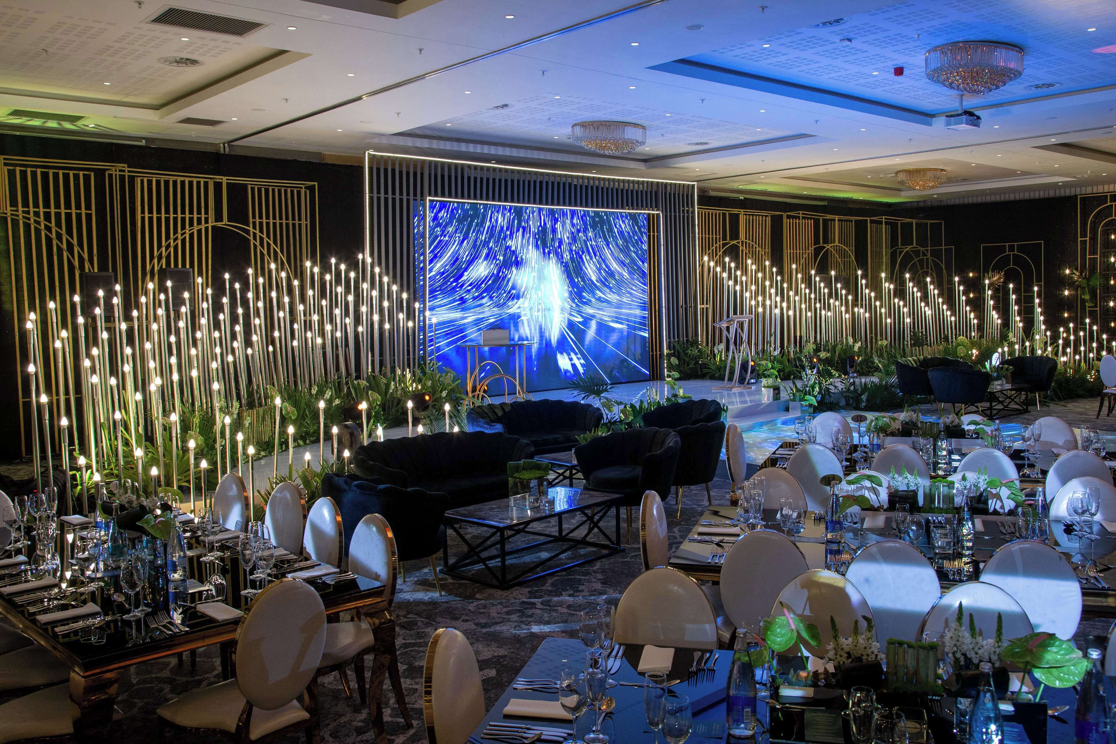 A formal dining area with tables set for a meal, a large screen displaying a blue abstract image, and decorative lighting.