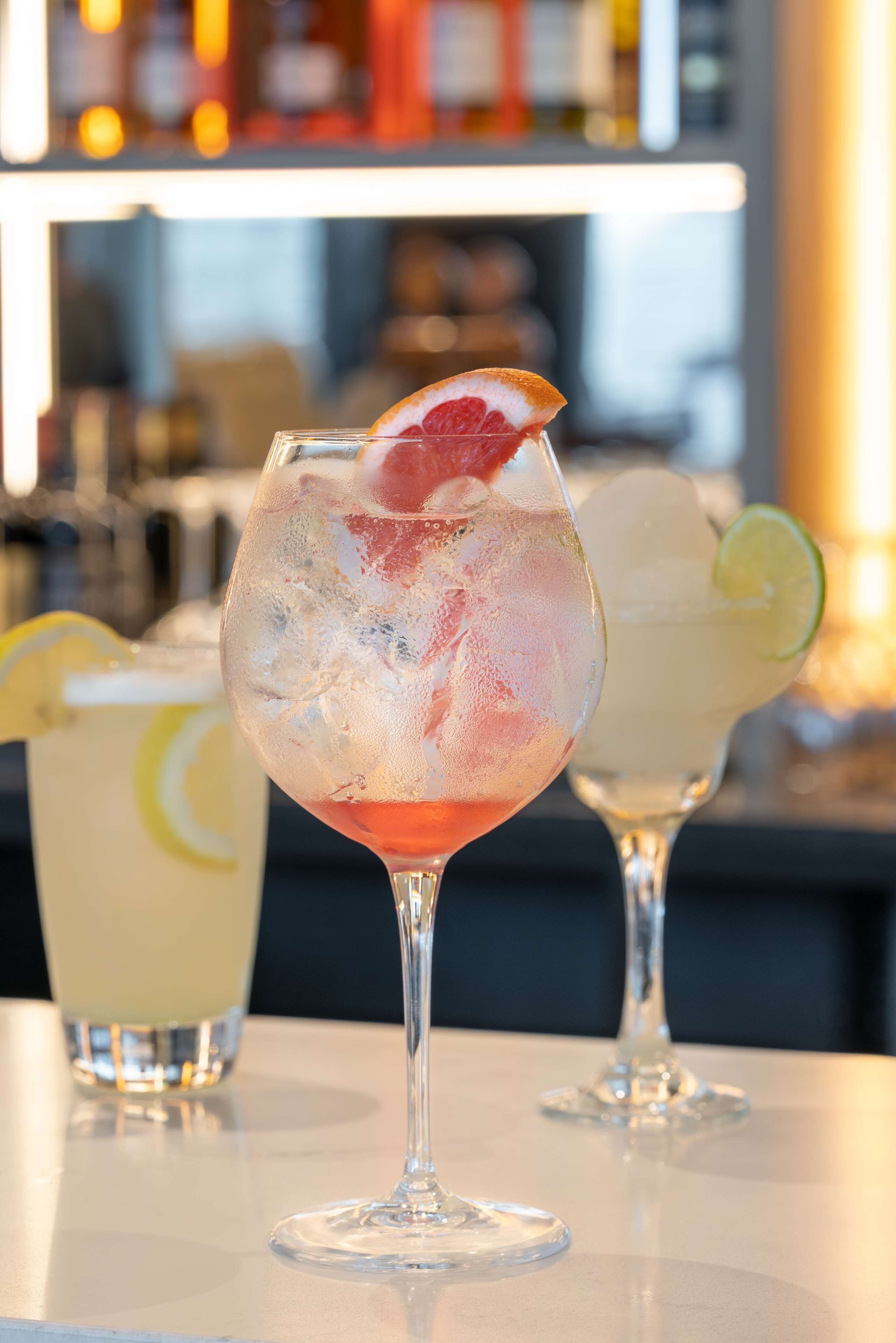 Three glasses of cocktails on a bar counter with a blurred background of bottles.