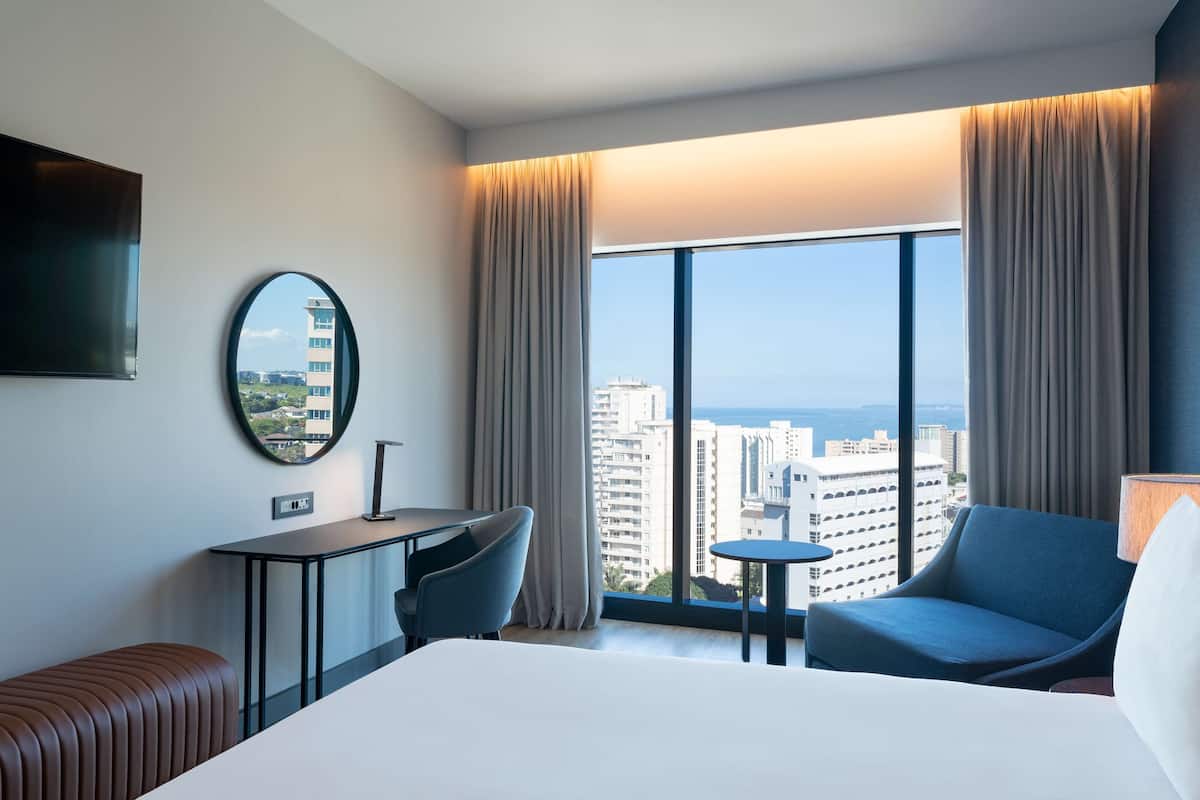 A hotel room with a bed, a desk, a chair, a small table, and a large window offering a city view.