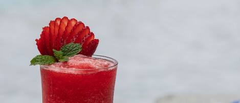 A glass of red beverage with a strawberry garnish on a white table by a beach.
