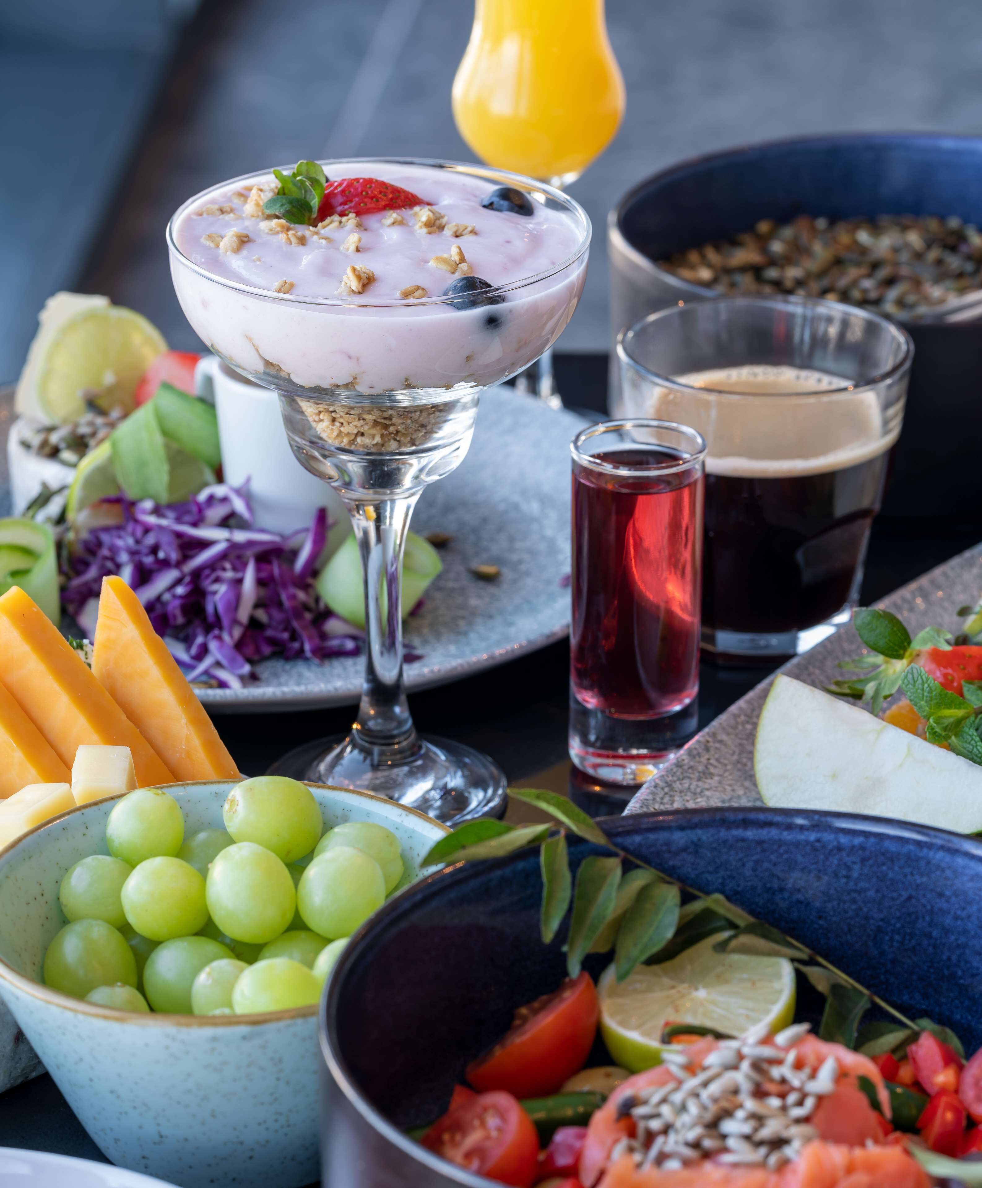 A table with a variety of food items including a glass of yogurt with granola and berries, a bowl of grapes, a plate of sliced vegetables, and a glass of dark beverage.