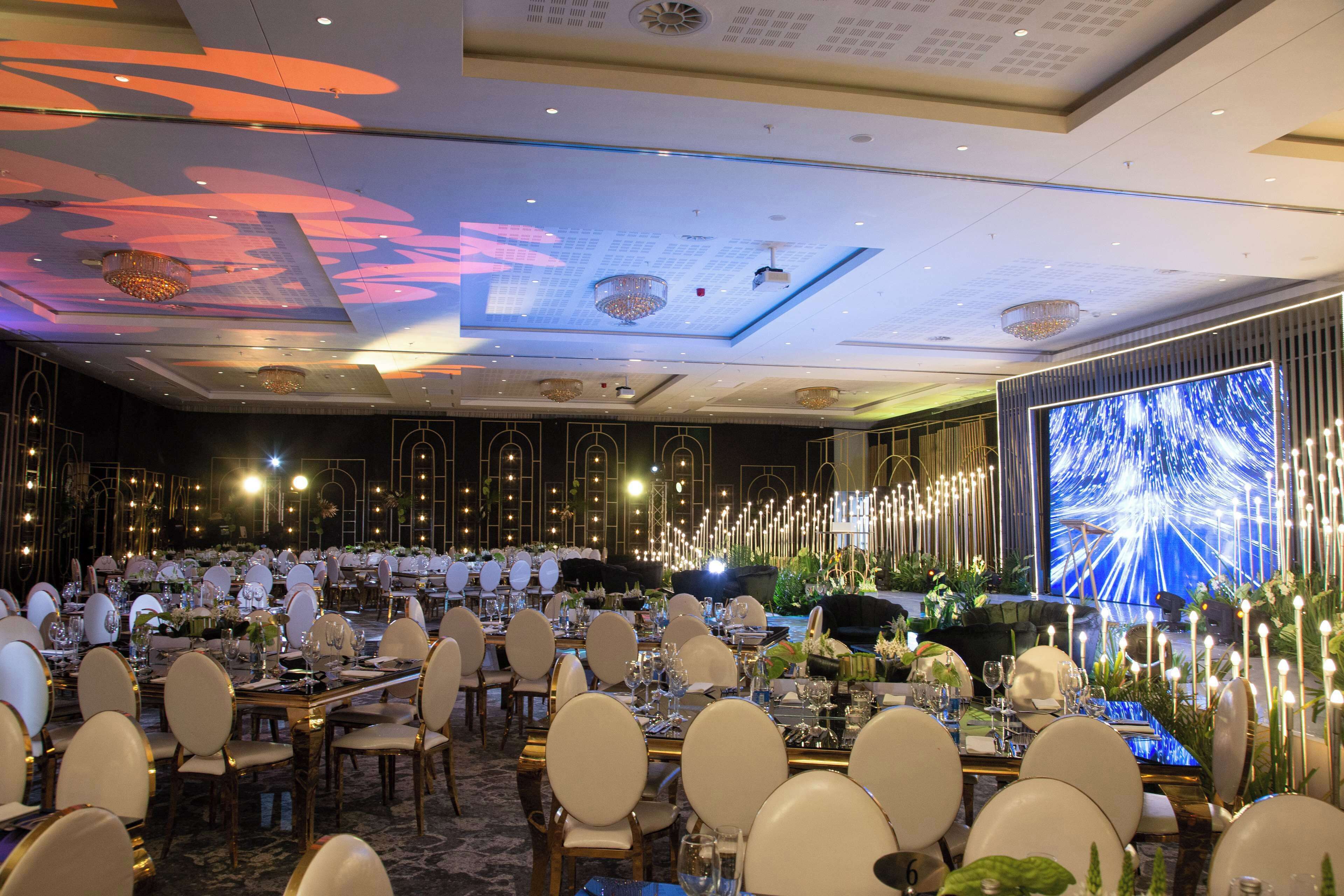 A banquet hall with round tables, chairs, and a large screen displaying a blue abstract image.