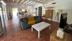 Amplia sala de estar con sofá, mesa de centro y televisor. Se observa una cocina al fondo y una escalera que sube a otro nivel.