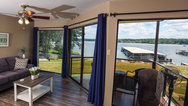 A room with a large sliding glass door leading to a lakeside view, a ceiling fan, a sofa, and a coffee table with a vase of flowers. - Lovely Lake Hamilton Condo, 1BR, Main Channel with an AMAZING VIEW! w/ boat slip (Hot Springs)