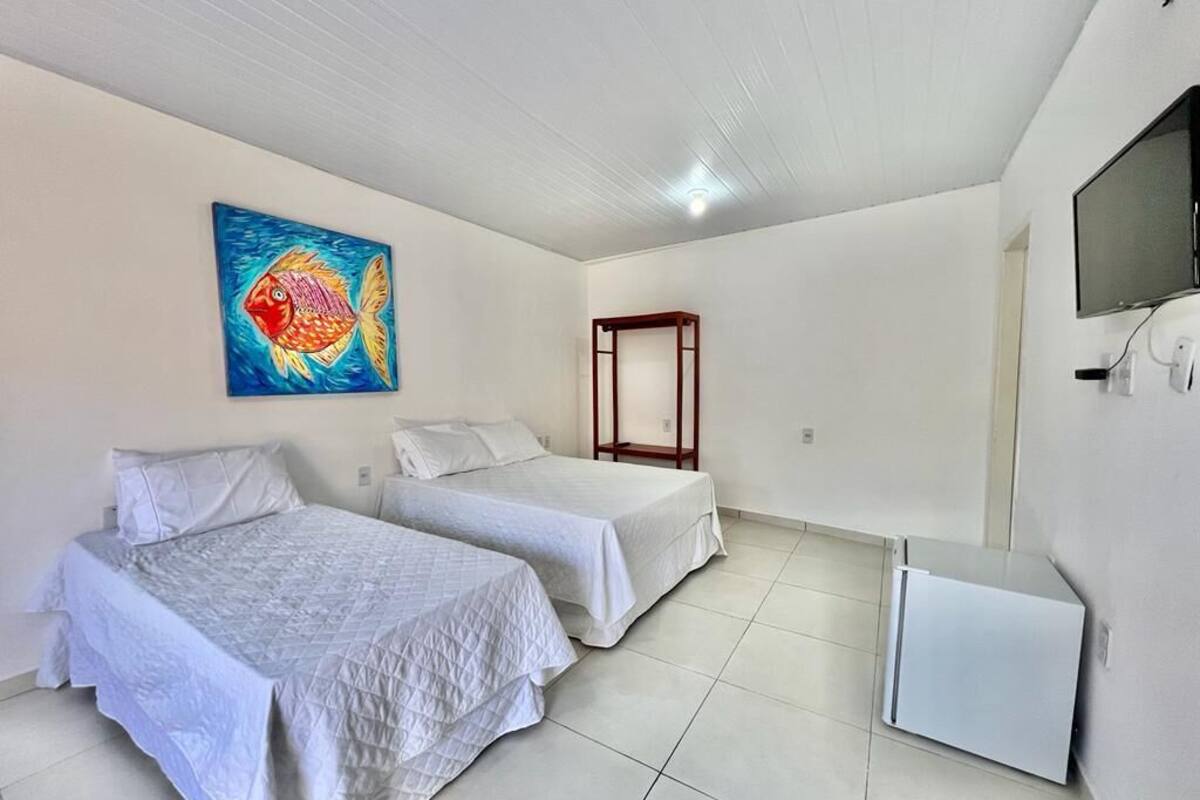 A room with two single beds, a small white cabinet, a wooden chair, a painting on the wall, and a television mounted on the wall.