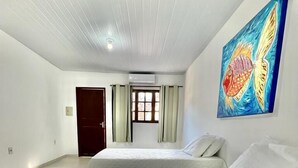 A spacious bedroom with a large bed, a window with curtains, an air conditioning unit, and a colorful fish painting on the wall. - Pousada Porto Henrris (Ipojuca)