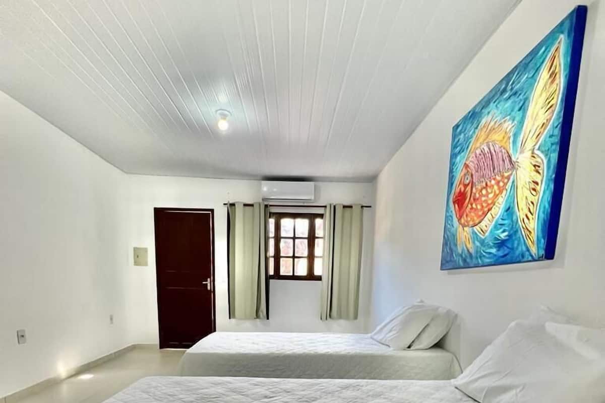 A spacious bedroom with a large bed, a window with curtains, an air conditioning unit, and a colorful fish painting on the wall.