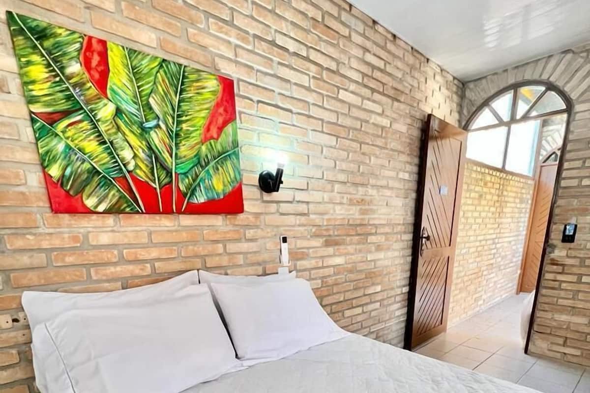 A modern hotel room with a brick wall, a large bed with white linens, a colorful tropical leaf painting, and a wooden door.