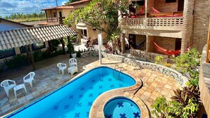 A swimming pool with a unique design, surrounded by a patio area with chairs and a building with a red roof. - Pousada Porto Henrris (Ipojuca)