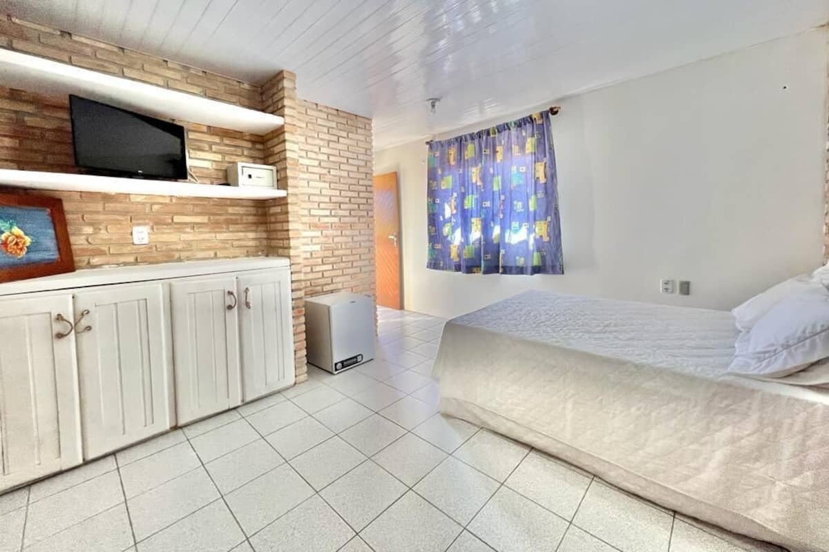 A bedroom with a bed, a TV on a shelf, a small cabinet, and a window with curtains.