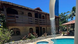 A swimming pool with a curved design, a water fountain, and a tiled patio area. - Pousada Porto Henrris (Ipojuca)