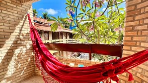A hammock in a room with a brick wall and a view of a building and trees. - Pousada Porto Henrris (Ipojuca)