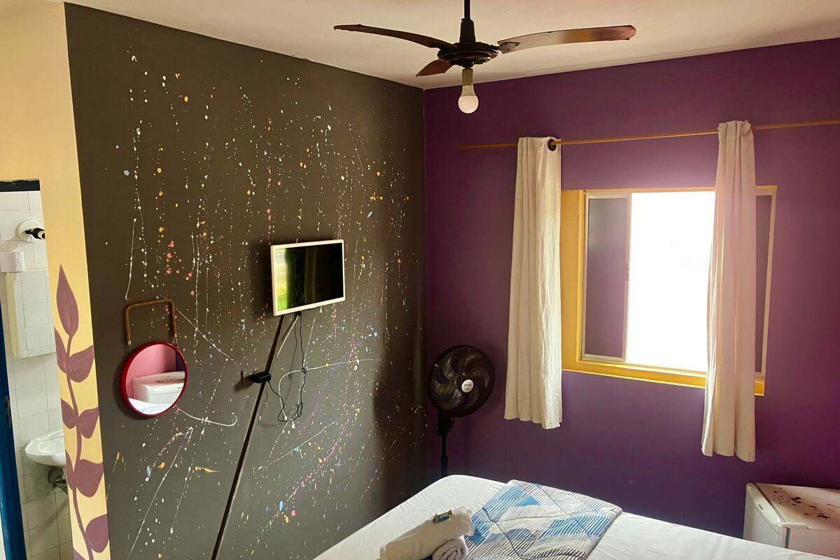 Une chambre à coucher comprenant un lit, un ventilateur de plafond, un téléviseur fixé au mur, un miroir, une fenêtre avec des rideaux et un mur orné d’un motif de feuilles.