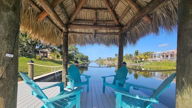 Een houten terras met turquoise stoelen, met uitzicht op een rustig wateroppervlak, een rieten dakgebouw en een helder zicht op een verre kustlijn met gebouwen.