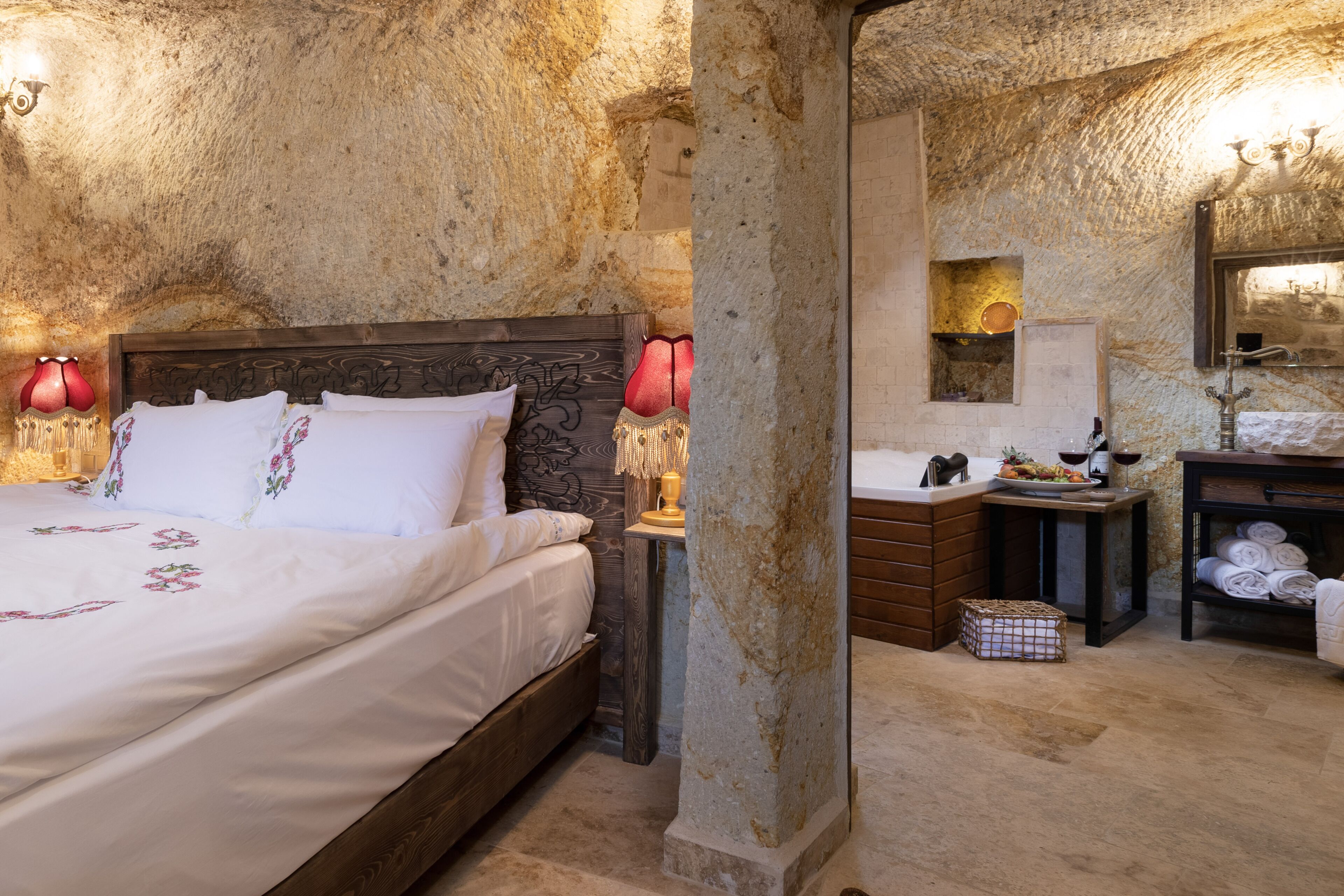 A bedroom with a large bed, a wooden headboard, a nightstand with a lamp, a stone wall, a small bathroom with a sink and mirror, and a shelf with towels.