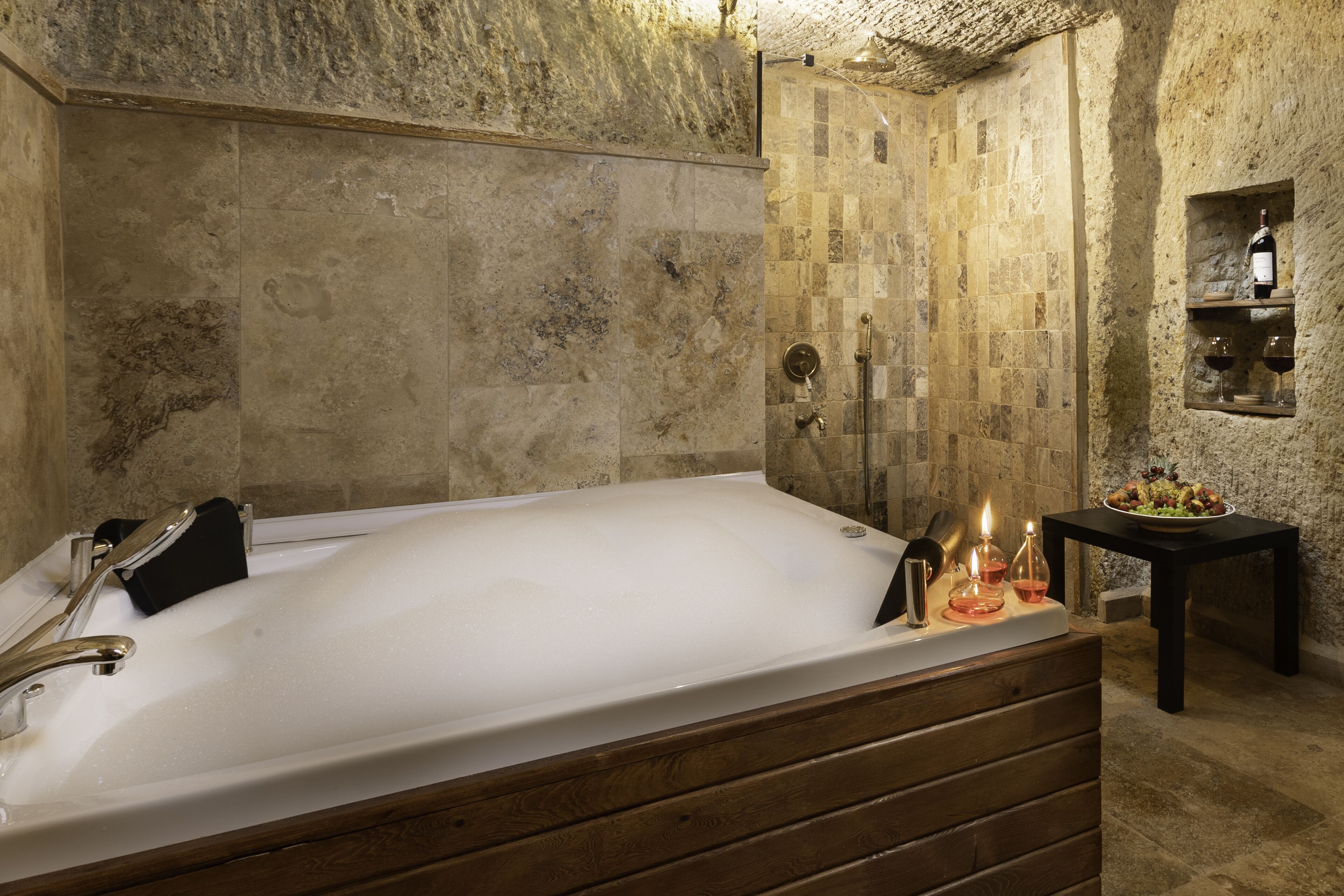 Eine große Badewanne mit Holzunterbau, ein wandmontiertes Regal mit einer Flasche und Gläsern sowie ein kleiner Tisch mit einer Obstplatte.