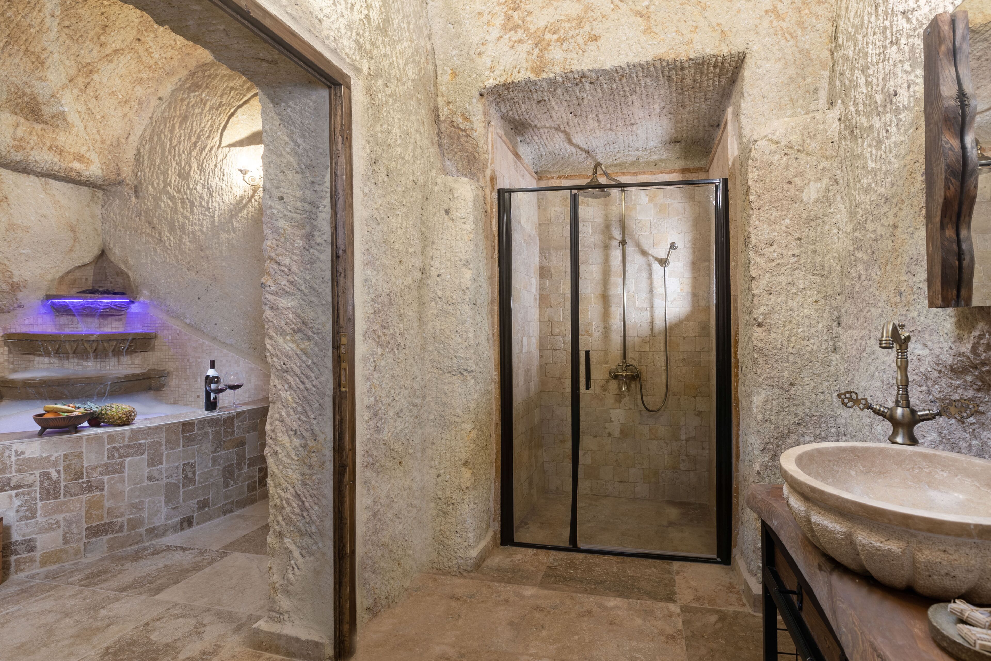 A stone-walled bathroom with a glass-enclosed shower, a sink with a vintage faucet, and a small alcove with a wine bottle and fruit.