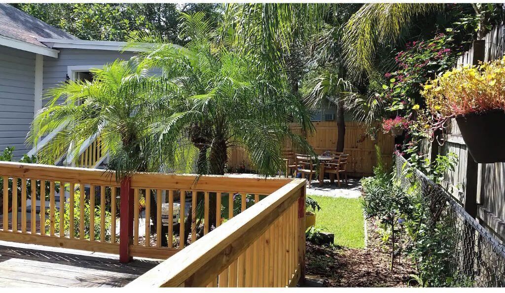 A wooden deck with a railing, a small patio area with a table and chairs, and a variety of plants including a palm tree and flowering plants.