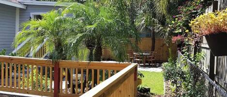 A wooden deck with a railing, a small patio area with a table and chairs, and a variety of plants including a palm tree and flowering plants.