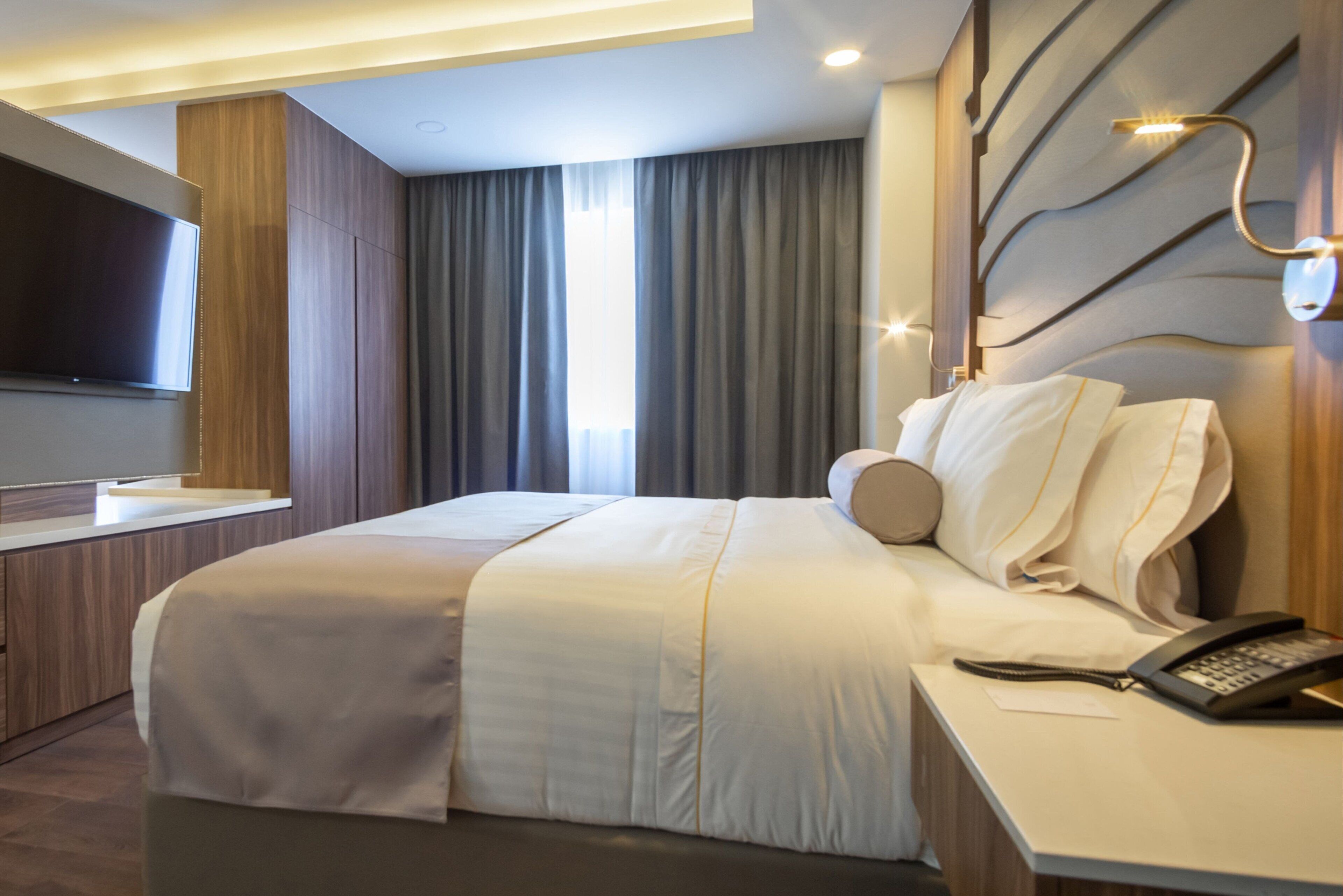 Ein modernes Hotelzimmer mit einem großen Bett, einem Nachttisch mit Telefon, einem an der Wand montierten Flachbildfernseher und einem Fenster mit Vorhängen.