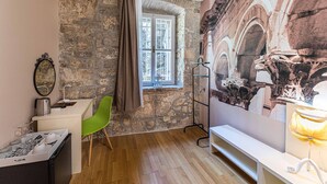 A room with a stone wall, a window with curtains, a desk with a chair, a bed, and a wall-mounted lamp. - Pjaca City Rooms (Split)